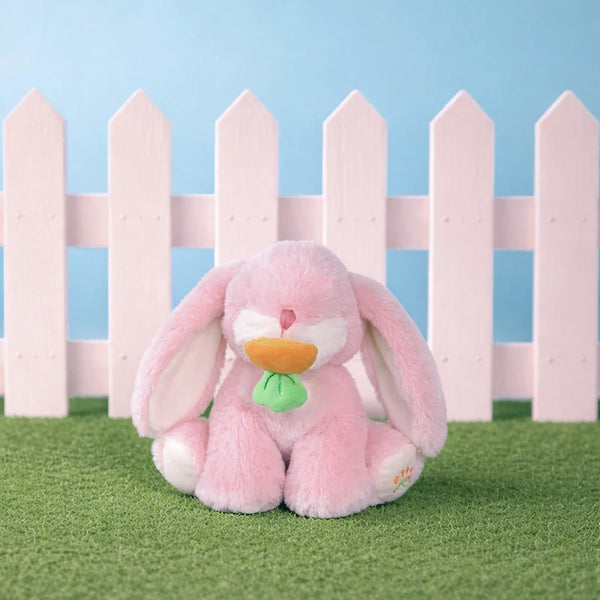 Pink plush bunny toy with a green leaf on grass in front of a white picket fence against a blue sky.