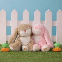 Two plush bunny toys, one brown and one pink, sitting on grass with a white picket fence and blue sky in the background.