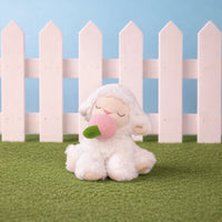 Plush toy sheep with a pink bubble gum sitting on grass with a white picket fence in the background