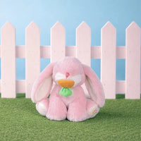 Pink plush bunny toy with a green leaf on grass in front of a white picket fence against a blue sky.
