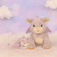 Two plush dragon toys on a soft, cloud-like surface with a pastel sky background.