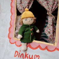 Small doll in a green coat and yellow hat peeking out from behind floral curtains with 'Dinkum' written below.