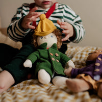Child holding a doll wearing a green coat and yellow hat on a checkered blanket.