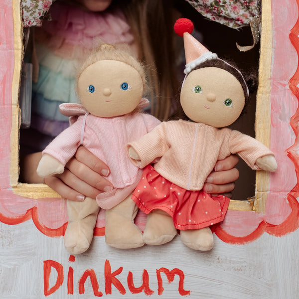 Two dolls held by a person with a 'Dinkum' label on a wooden surface.