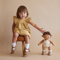 Plush baby doll with brown hair, beige romper, and brown shoes with a kid on a chair